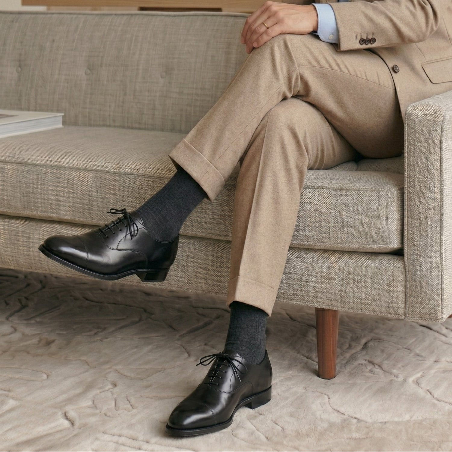 Person wearing a beige suit and black shoes sitting on a couch.
