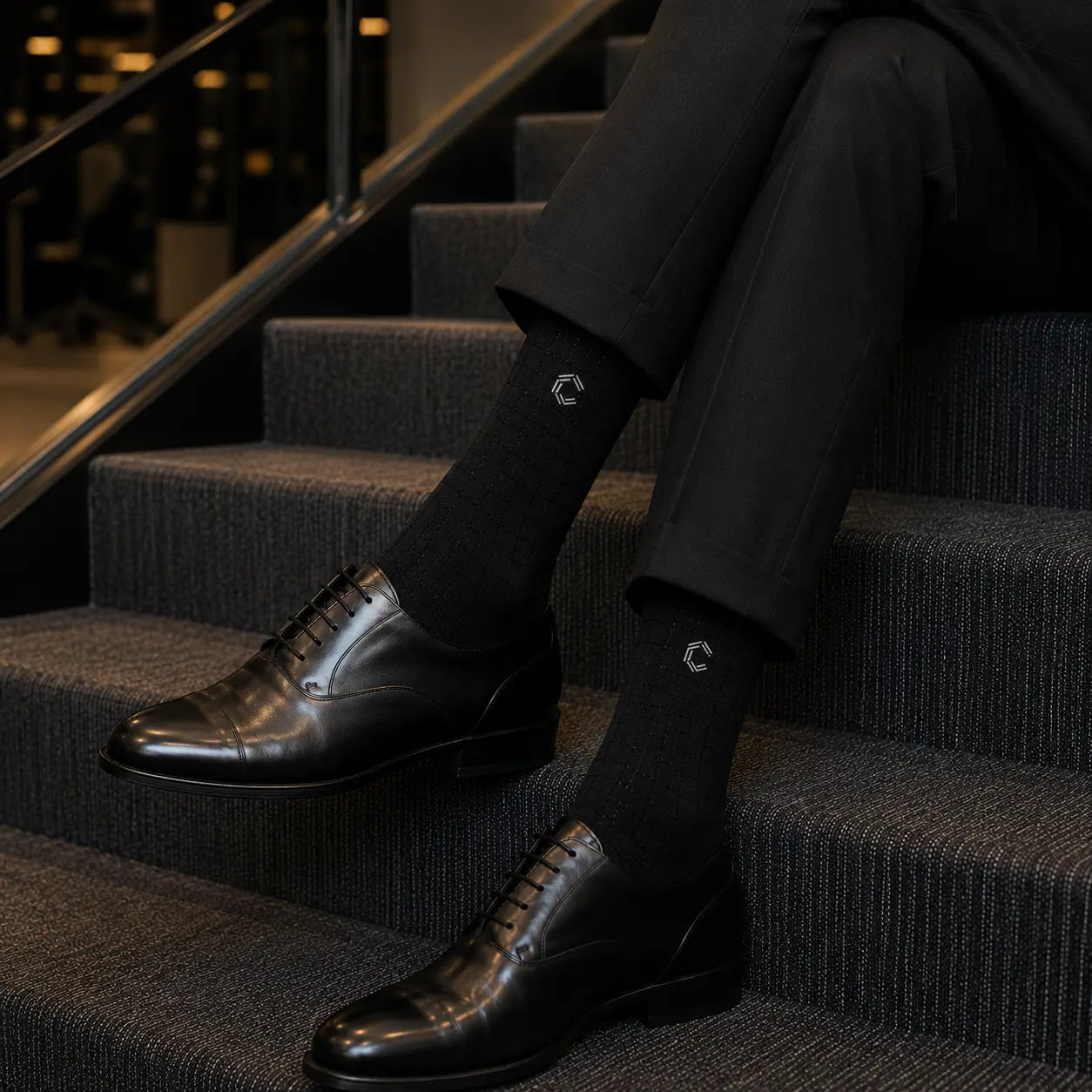 Person wearing black dress shoes and socks with a logo, sitting on stairs.